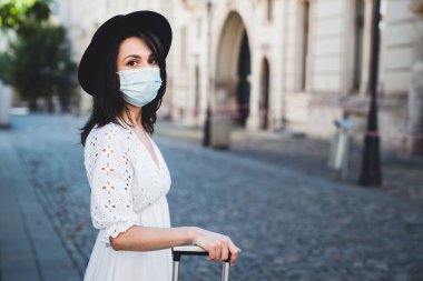 Koruyucu tıbbi maske takan genç ve güzel bir kadın seyahat çantası tutarken kameraya bakıyor. Beyaz elbiseli ve siyah şapkalı şık bir bayan turist salgın sırasında seyahat ediyor.
