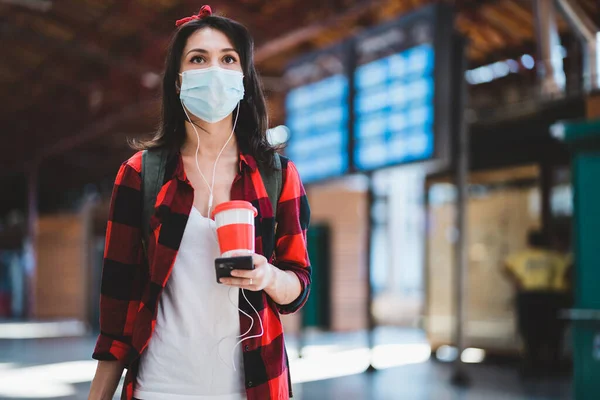 Life during covid-19 pandemic. Beautiful young woman waiting at bus or train station. Female wearing protective face mask and holding phone and cup of coffee travelling during coronavirus epidemic