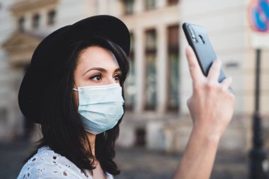 Covid-19 sırasında hayat. Koruyucu yüz maskesi takan genç bir kadın cep telefonuyla selfie çekiyor. Akıllı telefonuyla fotoğraf çeken şapkalı şık bir kadının portresi. Salgın sırasında sanal iletişim