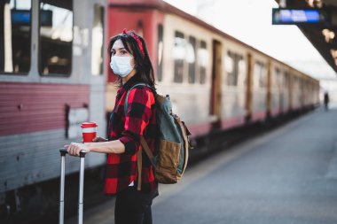 Tren istasyonunda koruyucu maskeli genç bir kadın bekliyor. Bir fincan kahve ve seyahat çantası taşıyan sırt çantalı bir turist. Salgın Seyahat Konsepti