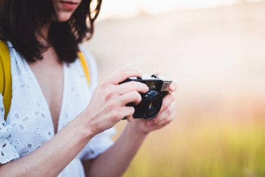 Genç bir kadın dışarıda klasik film kamerasıyla fotoğraf çekiyor. Tarz sahibi kadın sıcak gün batımına karşı retro kamera tutuyor.