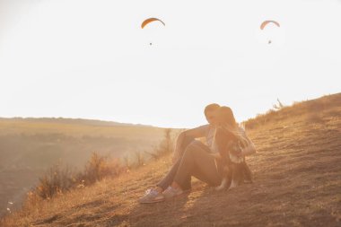 Birbirine aşık genç bir çift batan güneşin altında bir dağ yamacında oturuyor. Kız Schnauzer 'i kollarında tutuyor. Sarılırlar, havada süzülen paraşütçülere bakarlar. Yumuşak odaklı fotoğraf