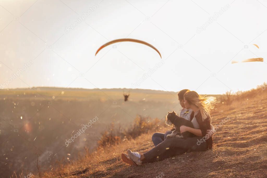 A young couple in love is sitting on a mountainside in the rays of the setting sun, the girl is holding Schnauzer in her arms. They hug,  look at soaring paragliders in the air. Photo with soft focus