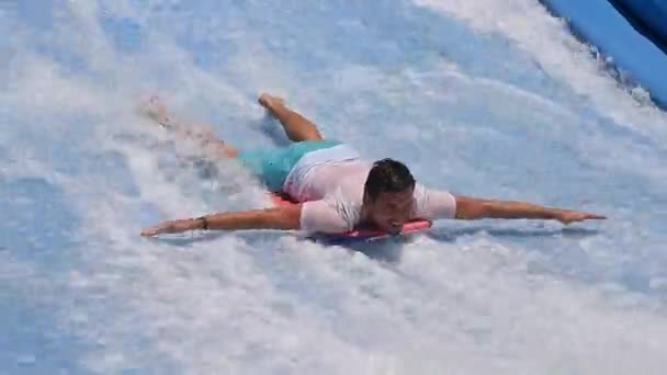 若い男が黒板に立って ウィンドサーフィンのために人工波に乗っている 白いTシャツと青いショートパンツの男がバランスを保とうとしていると彼の足に乗る トレーニングとシミュレーター — ストック動画