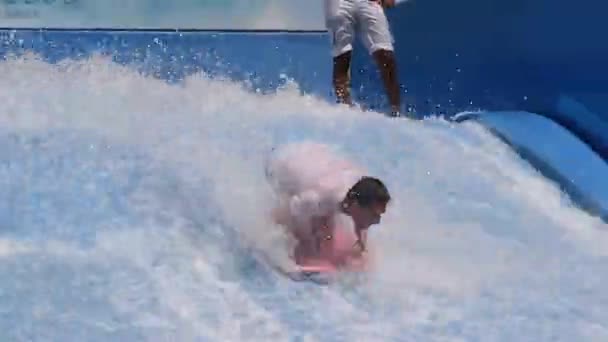 若い男が黒板に立って ウィンドサーフィンのために人工波に乗っている 白いTシャツと青いショートパンツの男がバランスを保とうとしていると彼の足に乗る トレーニングとシミュレーター — ストック動画