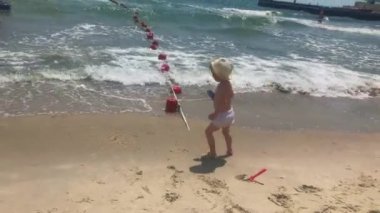 Hasır şapkalı ve beyaz şortlu küçük bir çocuk sahilde deniz kenarında oynuyor. Güzel bir kız sarı kumdaki beyaz köpüklü dalgalardan kaçar ve küreklerle oynar, kumdan kaleler yapar..