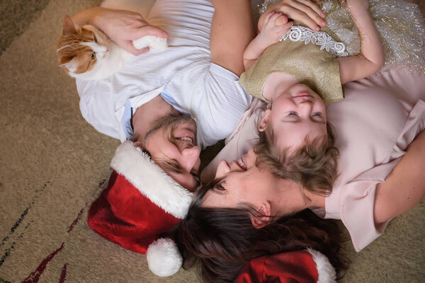 Beautiful family celebrate Christmas and New Year at home. On their parents' heads Christmas hat, they hug a little daughter and a ginger cat. Lying on the floor and laughing. 