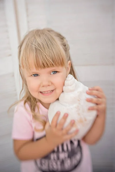 A beautiful little blonde girl with straight bangs with blue eyes and plays with seashells. Birthday party decorated with nautical decor. Happy cool baby girl plays on the seashore with her sister