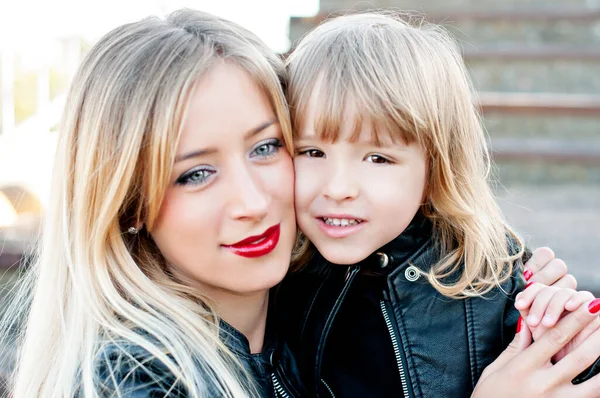 Beautiful mom blonde with red lipstick with a little daughter. A large portrait, looking at the camera, hugging and laughing. The theme of motherhood and pastime of mothers with children. Family look