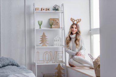 Güzel, modern, şık kıvırcık saçlı, gri örgü kazaklı, taytlı, boynuzlu bir kız. Noel sabahı pencerenin kenarındaki pencere pervazında oturuyor. Gülümsüyor, kameraya bakıyor.