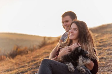 Aşık mutlu bir çift dağda siyah bir Schnauzer ile dinleniyor, yerde oturuyor, bir köpeğe sarılıyor, mavi gökyüzünde paraşütçülerin uçuşunu izliyor, gün batımını seyrediyor, bir köpekle seyahat ediyor.