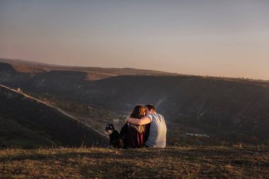 Aşık mutlu bir çift dağda siyah bir Schnauzer ile dinleniyor, yerde oturuyor, bir köpeğe sarılıyor, mavi gökyüzünde paraşütçülerin uçuşunu izliyor, gün batımını seyrediyor, bir köpekle seyahat ediyor.
