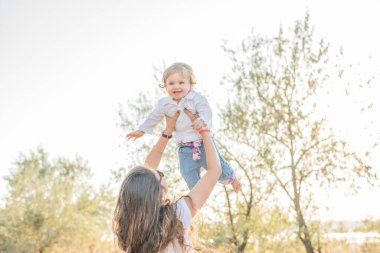 Uzun kıvırcık saçlı güzel esmer anne kucağına küçük bir kız alır gökyüzüne, kız güler, sevinir, kameraya bakar. Beyaz tişört ve kot pantolon giymişti. Anne!