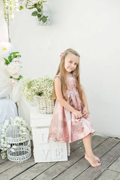 Beautiful little girl blonde with long hair with blue eyes. A close up portrait, looking at the camera, dressed in a powdery pink tender dress, standing by the bed with white chiffon canopies by sea