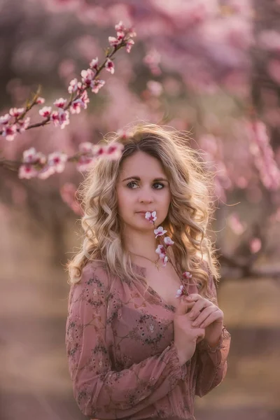 きれいな顔 自然なメイクで 美しい巻きブロンドの髪を持つ公開の新鮮な若い女の子は 開花庭に立って ピンクの花序を持つ小枝を保持しています 神秘的で 寛容で 敏感な女性は — ストック写真