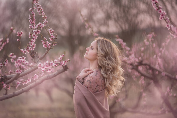 Tender fresh young girl with beautiful curly blonde hair, with a clean face, natural makeup, stands in blooming gardens, holds a twig with pink inflorescences. Mysterious, pensive, sensitive woman