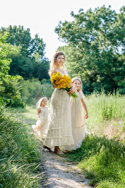家族の表情の同じ白い服を着た幸せな家族 美しいです母は立っています黄色の野花の花束周りに二人の小さな娘のブロンドの周りに陽気に実行され — ストック写真