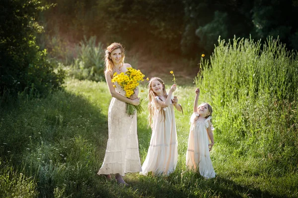 家族の表情の同じ白い服を着た幸せな家族 美しいです母は立っています黄色の野花の花束周りに二人の小さな娘のブロンドの周りに陽気に実行され — ストック写真