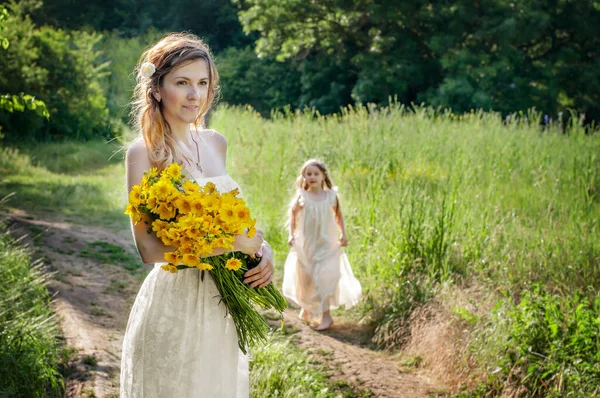 A happy family in identical white sundresses of a family look. A beautiful mother stands with a bouquet of yellow wildflowers around two little daughters of a blonde running around cheerfully and play