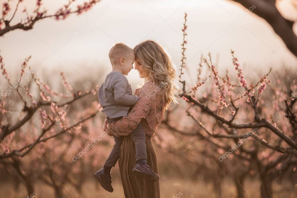 Beautiful young blonde mother with curly hair holding a little son in her arms in blooming gardens at sunset. Child care maternity concept. Mom throws the boy up, hugs, kisses, touches his forehead