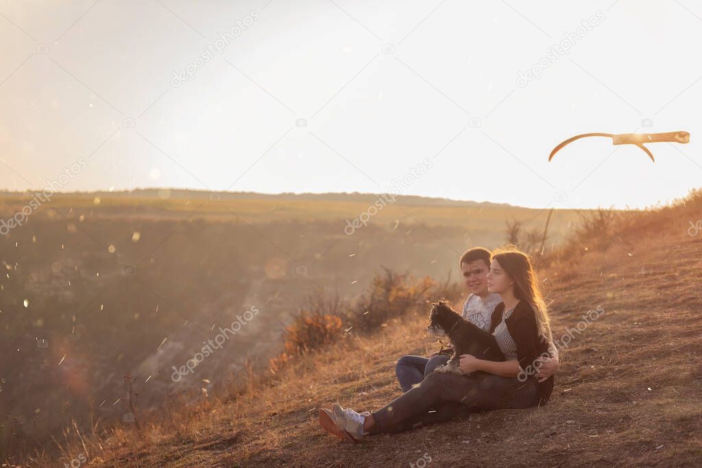 Happy couple in love are resting on the mountainside with a black Schnauzer, sitting on the ground, hugging a dog, watching bright  paragliders fly in the blue sky, admire the sunset, travel with a dog