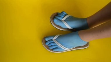 Kadın bacakları lastik mavi tıbbi eldiven, beyaz parmak arası terlik giymiş. Sarı üniformalı bir arka planda dans etmek. Virüs ve hastalıklardan korunmak için deniz kıyısında yaz tatili konsepti.