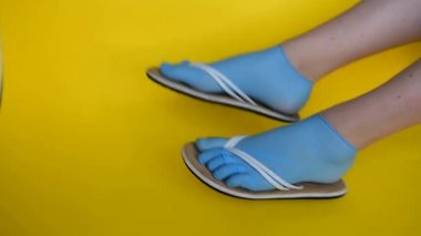 Kadın bacakları lastik mavi tıbbi eldiven, beyaz parmak arası terlik giymiş. Sarı üniformalı bir arka planda dans etmek. Virüs ve hastalıklardan korunmak için deniz kıyısında yaz tatili konsepti.