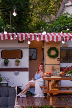 Kırmızı kıvırcık saçlı, mavi elbiseli, muhteşem bir gülümsemesi olan ince, güzel bir kız karavanının kapısında oturuyor. Şefkatli bir anne mutlu ailesiyle bir karavan arabasıyla seyahat eder. Bütçe tatili