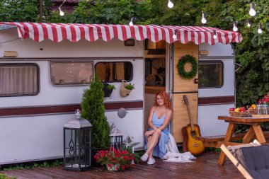 Kırmızı kıvırcık saçlı, mavi elbiseli, muhteşem bir gülümsemesi olan ince, güzel bir kız karavanının kapısında oturuyor. Şefkatli bir anne mutlu ailesiyle bir karavan arabasıyla seyahat eder. Bütçe tatili