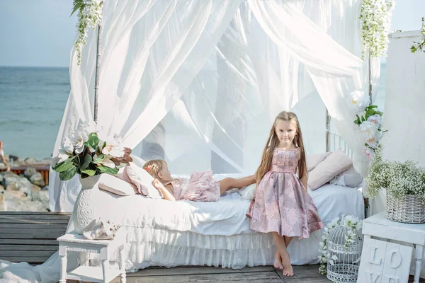 ピンクのドレスを着た2人の最高のブロンドの姉妹が一緒に遊んでいます 彼女たちは 藤の花が飾られた白いベッドの上に腰を下ろしている 幼い頃からの強い友情 — ストック写真
