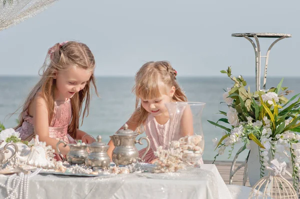 Beautiful blonde sisters girls in pink dresses are playing at the table in sea treasures behind the blue sea. Girlfriends are sorting through jewelry: beads, pearl earrings, shells, old dishes. Party