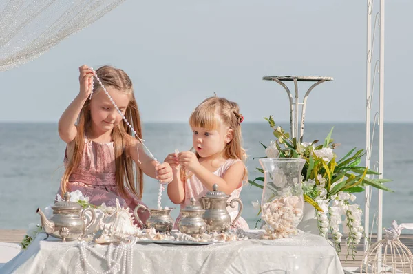 Beautiful blonde sisters girls in pink dresses are playing at the table in sea treasures behind the blue sea. Girlfriends are sorting through jewelry: beads, pearl earrings, shells, old dishes. Party