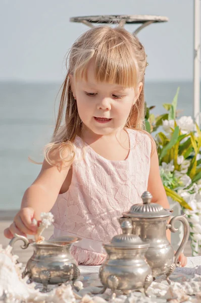Little cute blonde girl in a gentle pink dress plays with sea treasures at the table with a white tablecloth. A beautiful baby sorts out pearl beads, lays out shells, corals, examines old dishes.