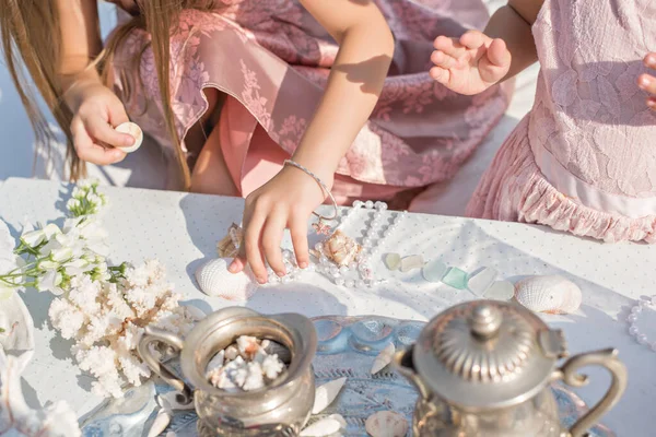 Little cute blonde girl in a gentle pink dress plays with sea treasures at the table with a white tablecloth. A beautiful baby sorts out pearl beads, lays out shells, corals, examines old dishes.