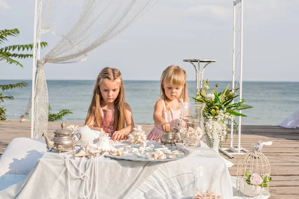 Beautiful blonde sisters girls in pink dresses are playing at the table in sea treasures behind the blue sea. Girlfriends are sorting through jewelry: beads, pearl earrings, shells, old dishes. Party