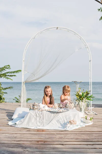 Beautiful blonde sisters girls in pink dresses are playing at the table in sea treasures behind the blue sea. Girlfriends are sorting through jewelry: beads, pearl earrings, shells, old dishes. Party