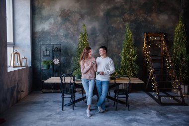 Sıcak kazaklar giyen sevgi dolu bir çift ellerinde turkuaz seramik bardaklar, kışın sıcak içecekler, Noel ışıklarının arka planında. Evde kış tatili atmosferi, Yeni Yıl kutlamaları.