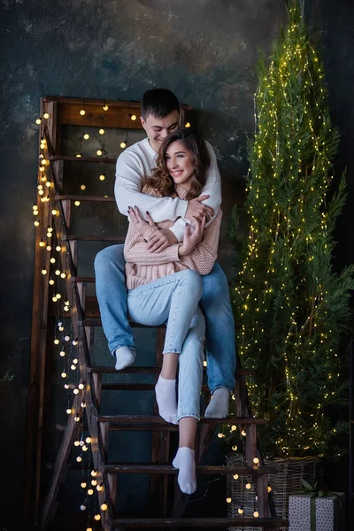 A young man with a beautiful girl in love are sitting on a wooden staircase decorated with Christmas lights. Lovers in warm winter sweaters hug, kiss, laugh. Harmonious relationship, New Year\'s date