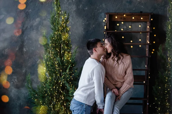 一个年轻人和一个漂亮的恋爱中的女孩坐在用圣诞彩灯装饰的木制楼梯上 穿着温暖冬季毛衣的情侣们拥抱 和谐的关系 新年的日子 — 图库照片