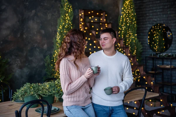 一对穿着保暖毛衣的恋人手里拿着两只绿松石瓷杯 在冬天喝着热饮 背景是圣诞灯 家庭寒假的气氛 新年庆祝活动 — 图库照片