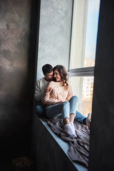 Happy couple in love in winter sweaters and jeans, sit on a wide windowsill by the window, hug. A young man kisses a beautiful curly brown-haired girl on the cheek. Date by the fireplace in a cozy home.