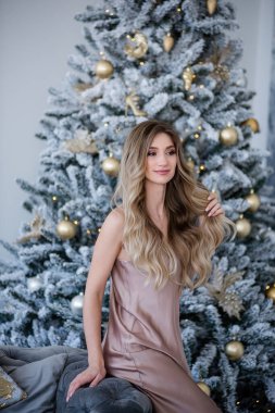 Şık, zarif, kendine güvenen, kıvırcık sarı saçlı, ipek tozlu, dar elbiseli, gri, modern bir kanepede oturuyor. Yeni yıl süslerinin ve hediyelerle dolu bir Noel ağacının önünde..