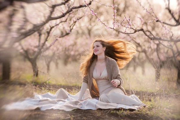 ベージュのボディコンドレスを着た美しい若い女性と茶色のニットカーディガンは 日没時に咲く桃の庭に鞭に座っています 彼女の赤みを帯びた長い髪は風になびく 思考の自由 日常からのリラックス — ストック写真