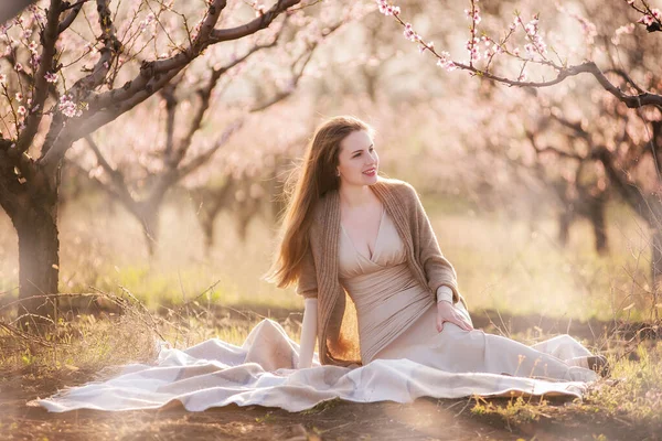 ベージュのボディコンドレスを着た美しい若い女性と茶色のニットカーディガンは 日没時に咲く桃の庭に鞭に座っています 彼女の赤みを帯びた長い髪は風になびく 思考の自由 日常からのリラックス — ストック写真