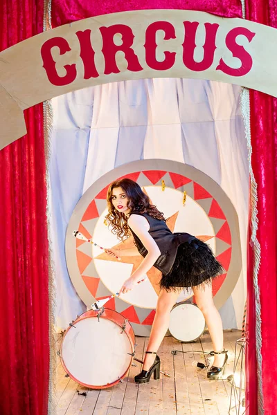 一个穿着洋娃娃服装的漂亮姑娘穿着黑色连衣裙拿着棍棒在鼓前打滚 站在木制地板上的红色马戏团的窗帘后面 魔术师的助手 万圣节演出 — 图库照片