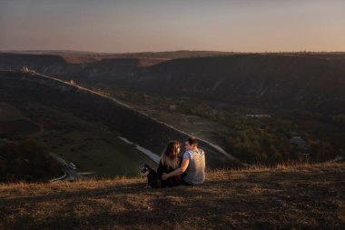 Kollarında Schnauzer köpeği olan sevgili bir çift gezgin bir kayanın üzerinde oturmuş birbirlerine sarılıyor ve nehrin gün batımını seyrediyorlar. Evcil hayvanlı mutlu bir ailenin gezisi 