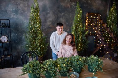 Genç bir çift, Noel için dekore edilmiş ahşap bir masada oturmuş sıcak içecekler içerken, küçük haydutlar yeşil çömleklere yerleştiriliyor, Noel ağaçları olan dokulu gri bir duvarın arka planında şenlik ışıkları yanıyor..