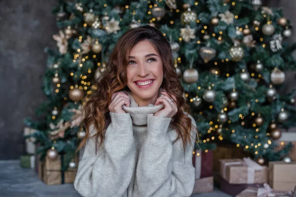 美しい巻き毛の女の子は 灰色のセーターの襟に包まれ 公然と笑顔 カメラを見るモデルのクローズアップポートレート おもちゃ 照明付きクリスマスツリーの背景 — ストック写真