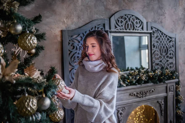 グレーのセーターを着た美しい巻き毛の女の子は 光のガーランドで飾られた大きな鏡でヴィンテージの暖炉のそばに立っています 近くにはクリスマスツリーがあり ギフトボックスが飾られています — ストック写真