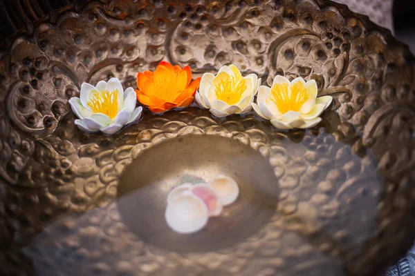 Beyaz ve turuncu nilüfer çiçekleri bakır bir kapta yüzerler. Kasenin altında deniz kabukları var. Spa, rahatlama konsepti. Manevi ifşanın, bilgeliğin, saflığın ve mükemmelliğin sembolü..